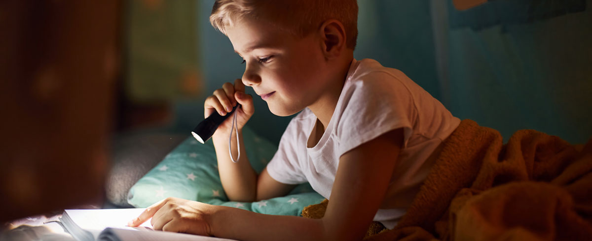 Un bambino che legge un libro con una torcia elettrica in mano mentre &egrave; nel letto con una coperta